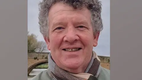 Karen Harvey Neil Harvey standing outside with a glimpse of a river and bridge behind him. He has short curling salt-and-pepper hair, has a scarf wrapped round his neck and is smiling broadly