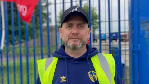 A man wearing a black cap with a white 'Unite' logo. He is also wearing a blue hoodie with a Leeds United logo, and a high-vis jacket. He is standing in front of a blue gate. The man has a grey beard.