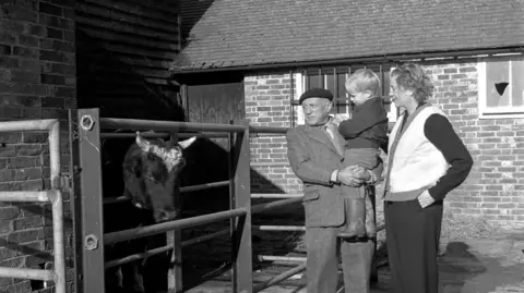 A man, Pablo Picasso, holding a child while stood next to a woman, Lee Miller. They are looking at a cow in a pen.