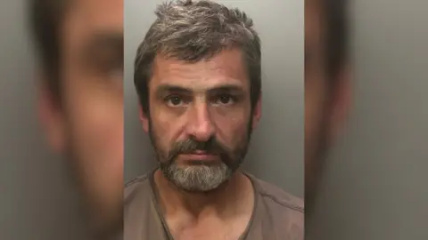 A man with greying hair and a beard in a police mug shot.
