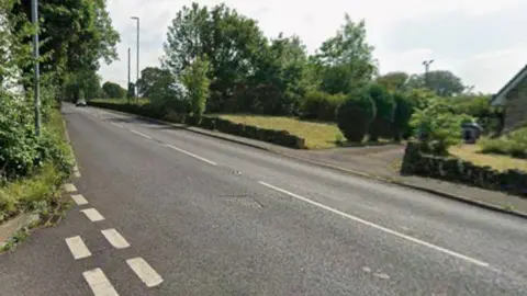 A quiet residential road in a suburban or semi-rural area on a bright day. The road runs diagonally from the bottom left corner toward the upper left, gently rising uphill. The surface of the road is dark grey tarmac with white painted markings, including dashed lines along the edge and a solid white line closer to the centre.