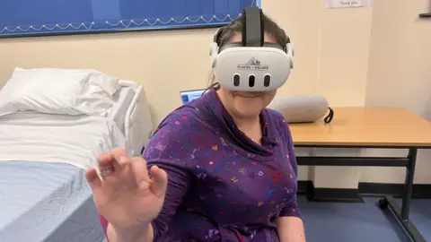 A woman wearing a virtual reality headset is pointing ahead of her. She is sitting next to a hospital bed in a training room at a hospital. 
