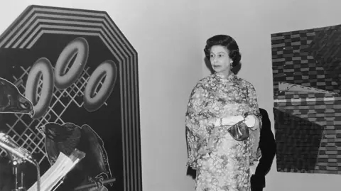 Courtesy of the Phillips family A black and white image of Queen Elizabeth in a1960s-style long flowery dress with a small handback. She is standing in front of a large piece of abstract art by the artist, comprising of two birds and large hoops. 