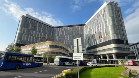 The front of the Queen Elizabeth University Hospital on a sunny day. The grey building has three tall rectangular sections and a curved beige building in front. There is a row of buses stopped outside and a car driving past.