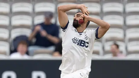 Zafar Gohar about to release the ball as he bowls for Middlesex, with white seats and a few spectators in the background