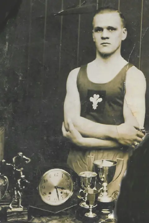 Drew family photograph Stan, in his 20s, poses defiantly before the camera with arms crossed. His vest bears the three feathers Welsh emblem. In the foreground can be seen several silver boxing trophies and an ornamental clock