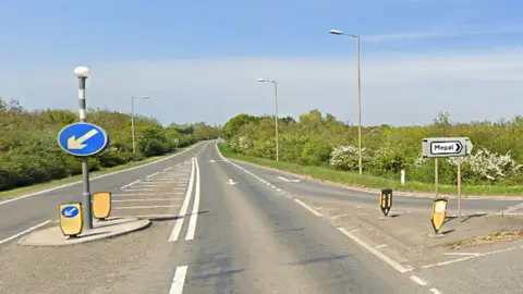 The A142 at the junction with Sutton Road, Mepal. It is a sunny day and there are no vehicles on the road. There is a sign pointing right for Mepal.