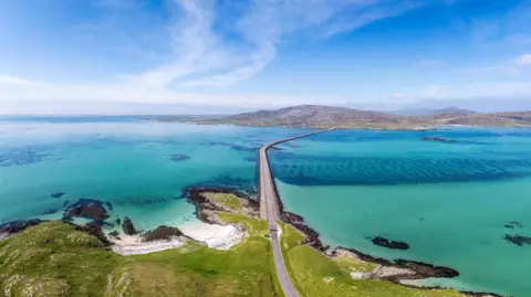 An aerial view of a causeway linking islands in the Western Isles. It is a beautiful day and the sky is blue with white wispy clouds. The sea is shallow and looks green.