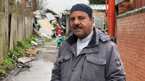 A man in a grey coat is standing in an alleyway and behind him is a large pile of rubbish which includes mattresses, wood and other furniture. 