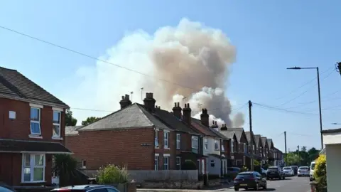 Jack Lott White smoke billowing above a row of bricked terraced houses, with cars parked on the road. The sky above is blue.