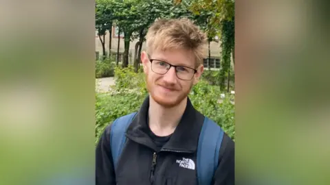 A man wearing glasses, a black fleece and a blue backpack. He is smiling at the camera.