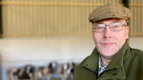 A man with a green coat and a brown tweed cap and wire framed glasses standing what appears to be the inside of a cattle shed