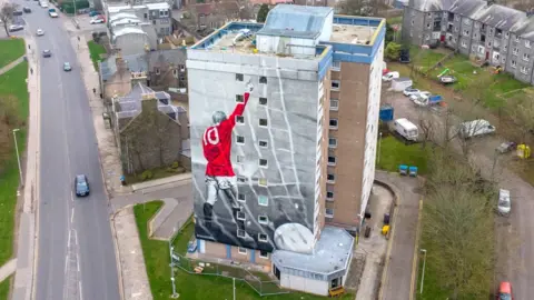 Painted image of the late footballer Denis Law in a red shirt, number 10 on back, arm raised, on the side of a high rise building, with Aberdeen in the background.