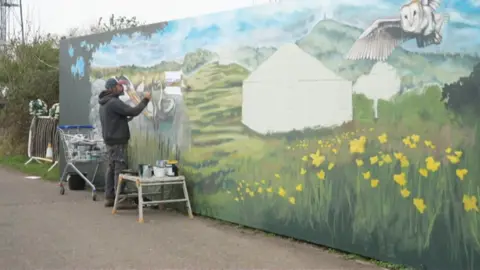 Artist Ady Szucik painting a commission on a hoarding. He has already painted Glastonbury Tor from a distance with yellow flowers and grass in the foreground of the mural. It also has a large owl in flight on it.