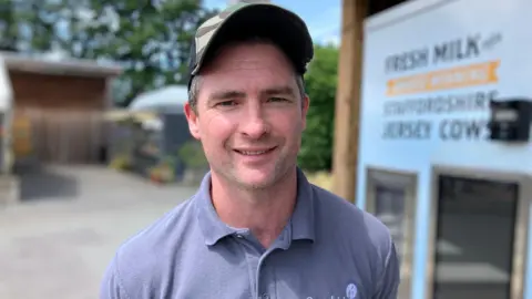 A man with a grey polo shirt and a camo cap on. he is looking at the camera smiling.