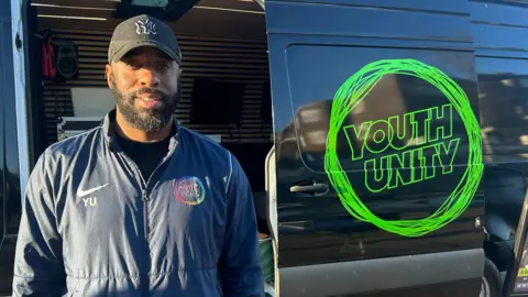 Youth Mentor Simeon Dennis is wearing a cap and standing in front of a van branded with the logo for youth group Youth Unity