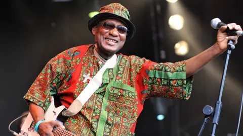 Getty Images Ebo Taylor in a colourful African shirt and hat and dark glasses. He is smiling and holding a microphone 
