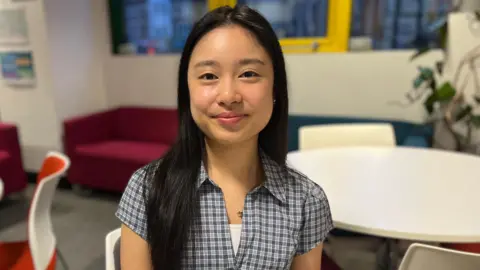 Kami smiling at the camera. She wears a checked shirt and has dark hair.