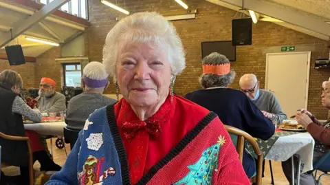 A close up of a woman with short white hair, smiling straight to the camera, sitting at a table in a community centre hall. She's wearing a blue and red Christmas woollen cardigan with a Christmas tree, a snowflake and a bear on it. Underneath she's wearing a red silky shirt and she has a red sequined bow tie around her neck. Behind her is a long table with people eating, some of them wearing paper crowns.