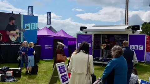 Stuart Howells/BBC The BBC Radio Suffolk area at a past Suffolk Show. A small stage area can be seen ahead as people watch someone singing into a microphone and playing a guitar. A large screen can be seen to the left.
