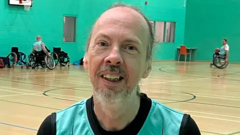 A man with short grey hair inside the sports hall, which has green walls. In the background are wheelchair basketball players using the court.
