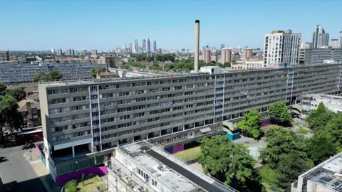 An aerial view of abandoned flats in south London. There are trees in bottom right of the frame and the city skyline can be seen in the distance against a blue sky.