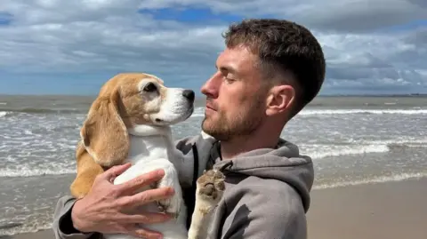 Aaron Ramsey Aaron Ramsey pictured on the beach with sea in background in grey hoodie holding the dog in his arms and looking at it, noses almost touching