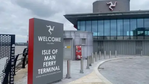 The outside of the ferry terminal, which has large glass windows in the second floor and a three-legs-of Mann symbol above. The is a grey sign that says Welcome - Isle of Man Ferry Terminal in English and Manx to the left and paving in front of it.