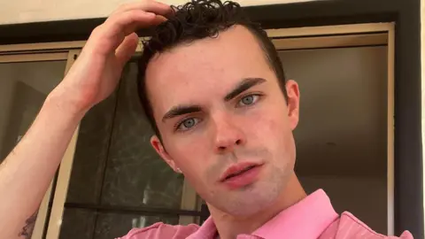 a young man with brown hair and blue eyes wearing a pink shirt.