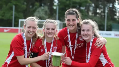 Getty Images Four female football players smile for the camera holding a silver trophy with a football pitch in the background