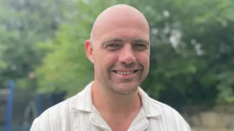 A bald man wearing a white shirt smiles at the camera. He appears to be outdoors
