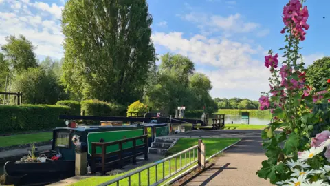 Rebekah A green and black canal barge sits in Northmoor Lock with green hedges and trees behind it. The foreground sees tall pink hollyhock flowers and white daisies. The sky is sunny, blue and dappled white clouds.