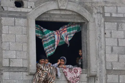 Mulheres e crianças da Reuters espiam pelas janelas de um prédio de pedra. O carpete está espalhado pela janela e pendurado no teto.