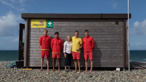 RNLI Joanna standing in front of the lifeboat station at Newgale Beach with Matty and three other lifeguards. Joanna is in the middle and next to her, on the right, is Matty. The other lifeguards are wearing their RNLI uniform - a red long sleeve top, yellow t-shirts underneath the long sleeve tops and red shorts.
