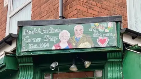 A green hand-painted sign above a door which says Derek and Pauline's corner shop and so much more. It features a painting of an elderly couple smiling. In the portrait she has white hair and is wearing glasses, a pink gilet and blue top. He is wearing a khaki overcoat and a blue polo shirt.