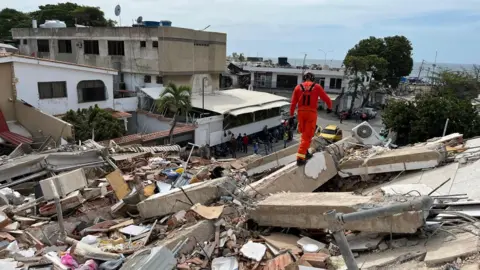 UK ISAR A search and rescue team member in an orange overall walks over an enormous pile of rubble.