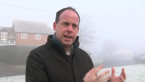 Steve Humphrey/BBC MP Greg Smith gestures with his hands to express his feelings against the biogas plant. He stands on a grass verge near a road, with a house and hedge behind him, on a very foggy day. He wears a white checked shirt, a teal jumper and a cushioned black jacket.