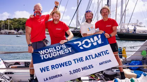 Four men smiling holding a sign which says "World's toughest Row. We rowed the Atlantic. La Gomera - 3,000 miles - Antigua".