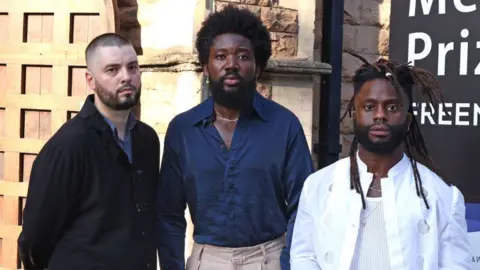 Getty Images Graham Hastings, Alloysious Massaquoi and Kayus Bankole of Young Fathers attends The Mercury Prize 2023 awards show at Eventim Apollo on September 07, 2023 in London. Graham is a white man in his 30s with shaved dark hair and a short dark beard. He wears a black jacket over a blue shirt. Alloysious stands beside him - he is a slightly taller black man also in his 30s. He has afro hair and a beard about two inches long. He wears a blue shirt with the top two buttons undone, revealing his chest and a gold chain necklace. Beside him is Kayus, who is the shortest of the three artists. He is a black man with his hair styled in dreads and a short beard. He wears a white shirt open over a white vest with three necklace chains and a nose ring