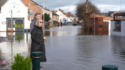 A man and a woman, both in winter coats, survey the flood waters in Station Road, Quorn, which is around ankle depth. Ripples can be seen in the water and the Monk restaurant partly submerged in the background.
