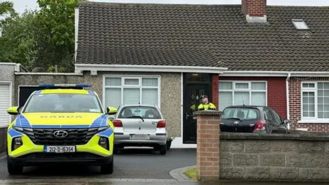 A police officer is seen standing outside the door of a house where two cars are parked along with a police jeep.