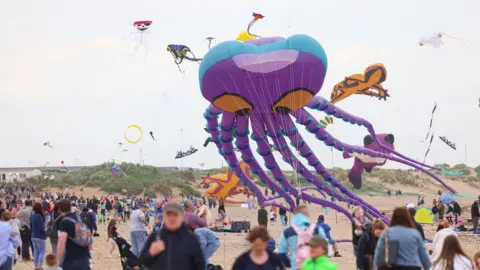 A large purple kite in the shape of an octopus being flown at Redcar's beach. It has large orange eyes. A number of other kites can be seen in the air in the background including an orange lizard. Dozens of people are watching on.