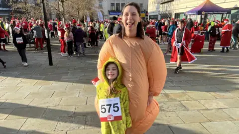 Lauren and her daughter laugh as their picture is taken. Lauren wears an inflatable turkey costume and her daughter wears a yellow onesie with 'the grinch' written on it. Behind them lots of people in santa costumes are stood around in the square. 
