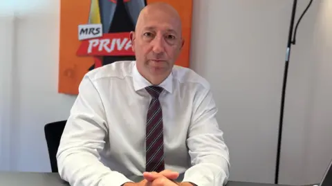 Paul Vane sitting at a desk with his hands clasped. He has a serious expression, and is wearing a white shirt and stripey tie. He is bald. There are campaign posters visible in the background.