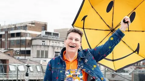 Sunny Turner grins as she holds aloft a yellow umbrella with a smiley face on an urban rooftop with high-rise buildings behind. She wears a a blue denim jacket and orange shirt under bright dungarees.