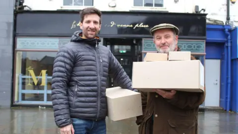 Durham University/The Auckland Project John Castling (left) and Prof Chris Gerrard (right) are smiling while standing in front of the windows of the Mrs M venue in Bishop Auckland. They are each holding cardboard boxes. Ms Castling is wearing jeans and a black puffer jacket. he has a beard and short brown hair. Prof Gerrard is wearing a khaki bakerboy cap and a brown coat. He has white beard. 