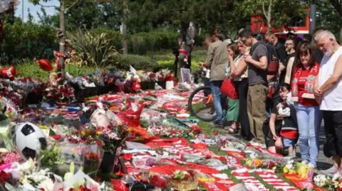 A crowd of fans pay respects at tributes to Jota. One woman has a hand over her mouth. A man in a white vest bows his head to reflect. 