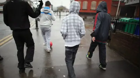Four young boys walk down a street in the rain with two carrying a football. They have their backs to the camera and their hoods up. They are walking past buildings.