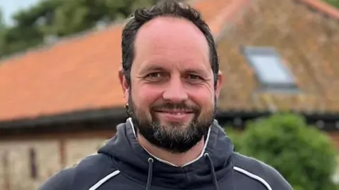 Family photo Rhys Jenkins who has dark hair, beard and moustache, wearing a navy and white hoody and smiling at the camera