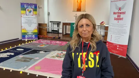 Hayley Smith with long blond hair wearing a navy hoodie with the word Test in yellow writing and a HIV red ribbon and red lanyard standing inside St Andrw's Church with the panel of the memorial quilt on show on the floor behind her